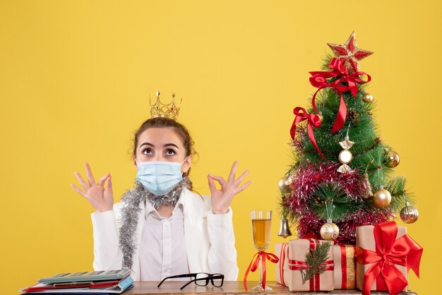 크리스마스 트리와 선물 상자와 노란색 배경에 멸균 마스크에 앉아 전면보기 여성 의사
