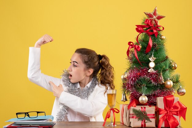 Front view female doctor sitting behind table flexing on yellow background with christmas tree and gift boxes