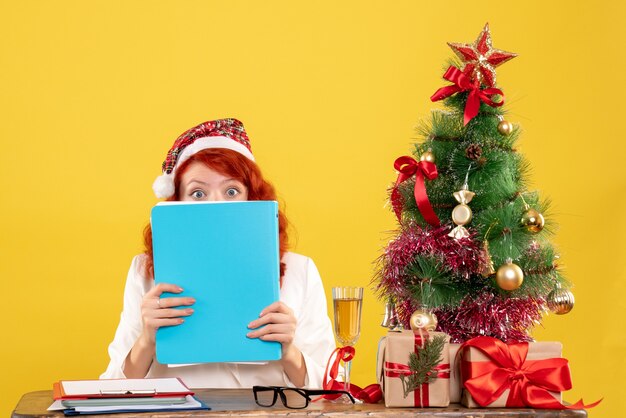 테이블 뒤에 앉아 크리스마스 트리와 선물 상자와 노란색 배경에 문서를 들고 전면보기 여성 의사