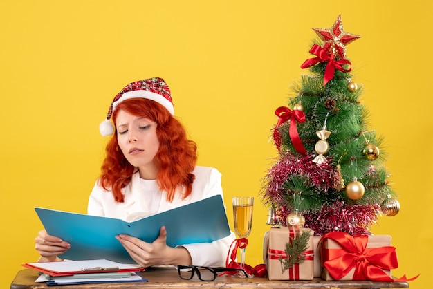 Free photo front view female doctor sitting behind table and holding documents on yellow background with christmas tree