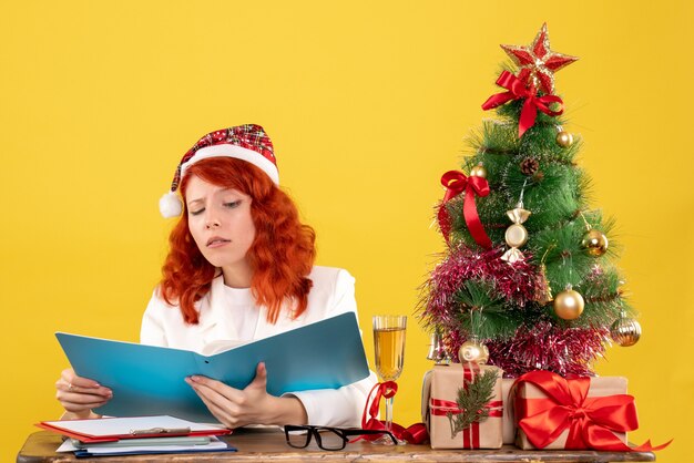 Front view female doctor sitting behind table and holding documents on yellow background with christmas tree