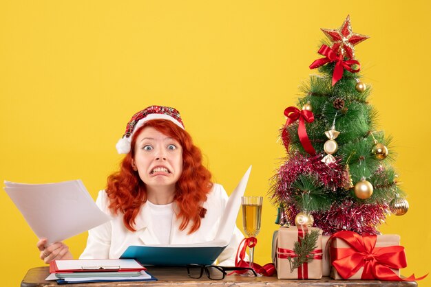 Free photo front view female doctor sitting behind table and reading documents on a yellow background with christmas tree and gift boxes