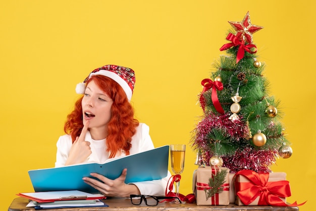 Free photo front view female doctor sitting behind table and reading documents on yellow background with christmas tree and gift boxes