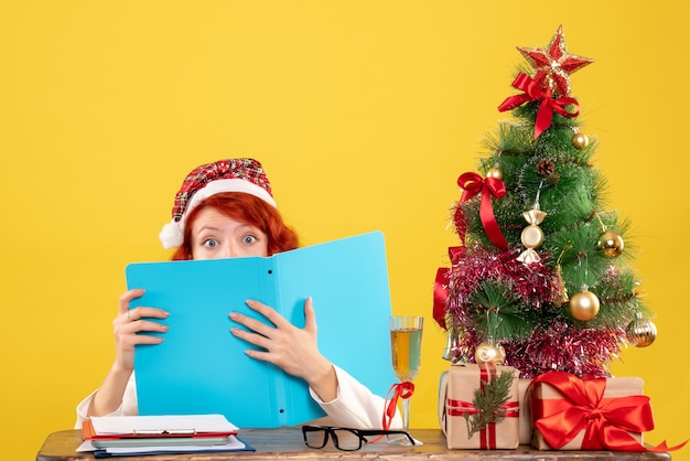 Front view female doctor sitting behind table reading documents on yellow background with christmas tree and gift boxes