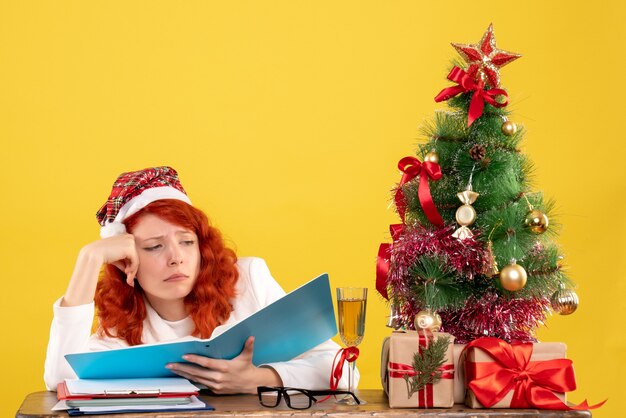 クリスマスツリーとギフトボックスと黄色の机の上のドキュメントを読んでテーブルの後ろに座っている正面図の女性医師