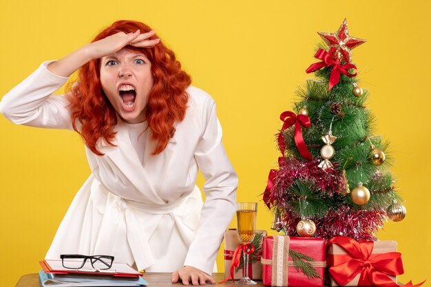 Front view female doctor sitting behind table with christmas presents feeling angry on yellow background