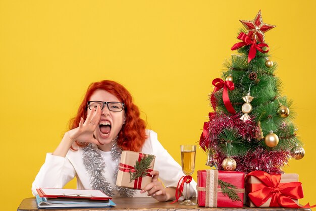黄色の背景で叫んでクリスマスプレゼントとテーブルの後ろに座っている正面図の女性医師