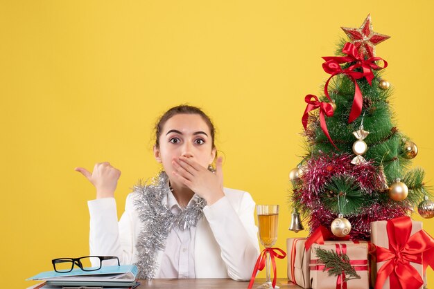 クリスマスプレゼントとクリスマスツリーとギフトボックスと黄色の背景の木とテーブルの後ろに座っている正面図女性医師