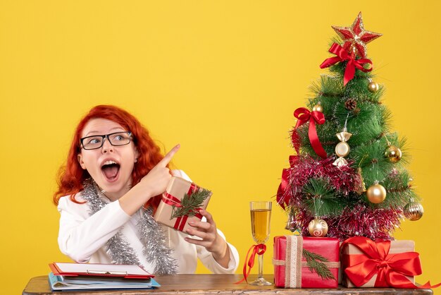 Front view female doctor sitting behind table with christmas presents on a yellow background