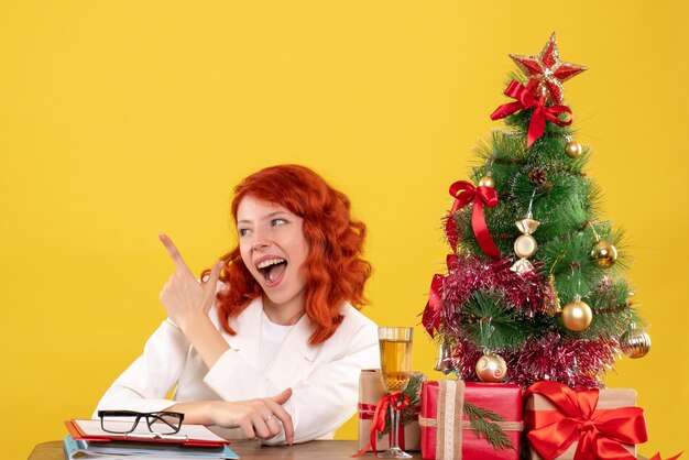 Вид спереди женщина-врач сидит за столом с рождественскими подарками на желтом фоне