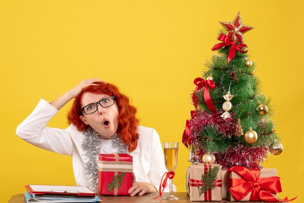 Front view female doctor sitting behind table with christmas presents on a yellow background