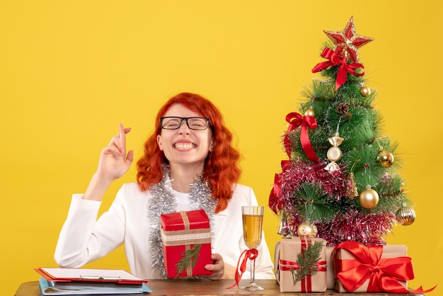 Medico femminile di vista frontale che si siede dietro la tavola con i regali di natale su fondo giallo