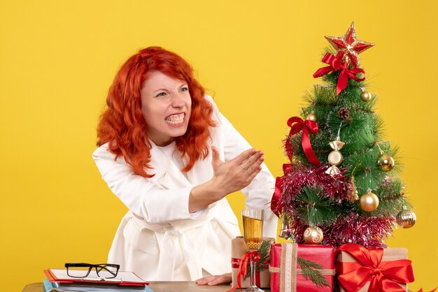 Front view female doctor sitting behind table with christmas presents on a yellow background