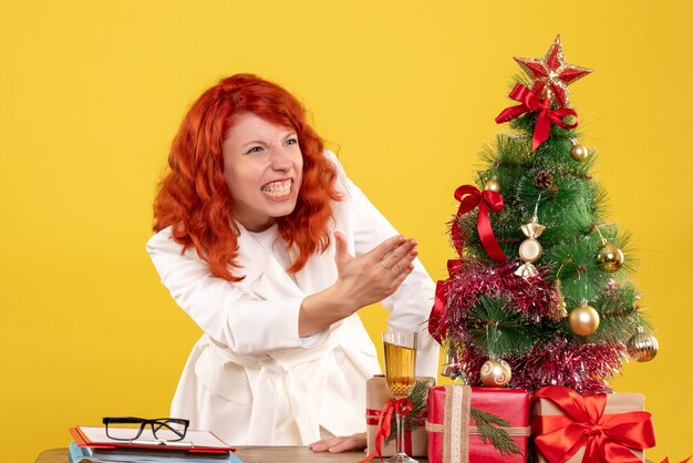 Вид спереди женщина-врач сидит за столом с рождественскими подарками на желтом фоне
