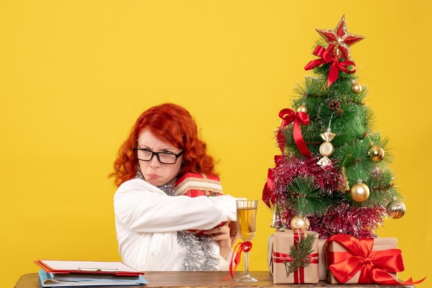 Front view female doctor sitting behind table with christmas presents on the yellow background
