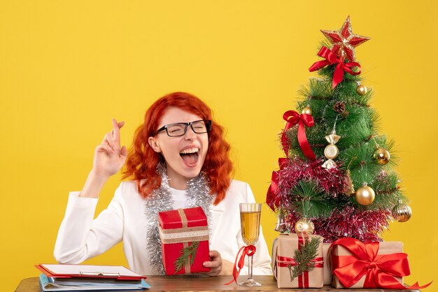 Front view female doctor sitting behind table with christmas presents on the yellow background