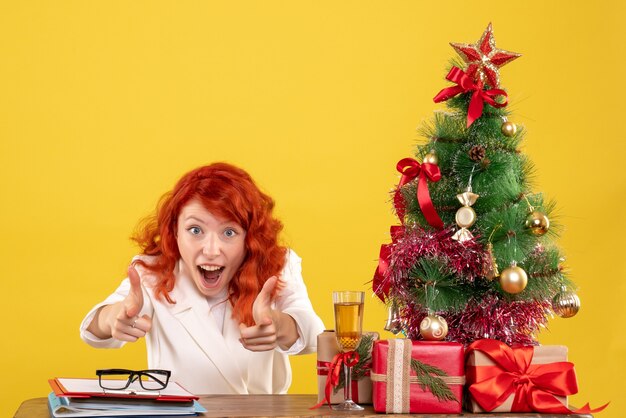 Front view female doctor sitting behind table with christmas presents on the yellow background