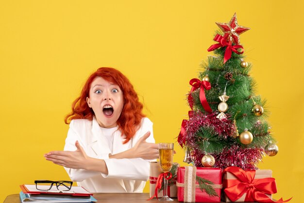 Front view female doctor sitting behind table with christmas presents on the yellow background