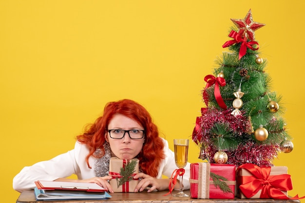 Foto gratuita medico femminile di vista frontale che si siede dietro la tavola con i regali di natale su fondo giallo