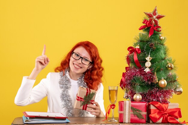 Front view female doctor sitting behind table with christmas presents on yellow background