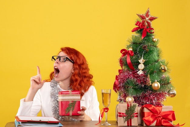 Front view female doctor sitting behind table with christmas presents on yellow background