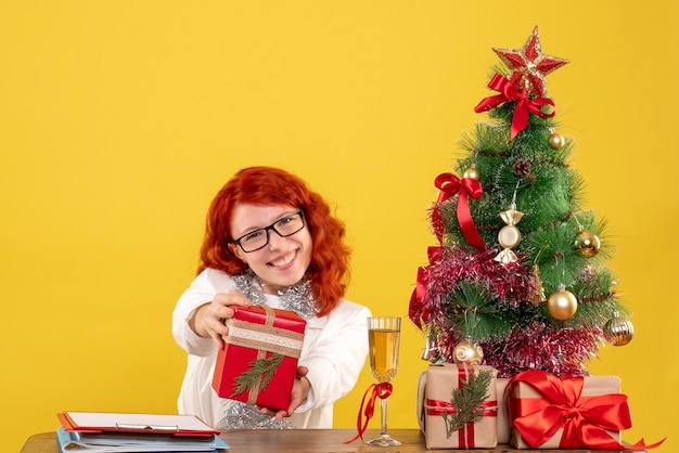 Front view female doctor sitting behind table with christmas presents on yellow background