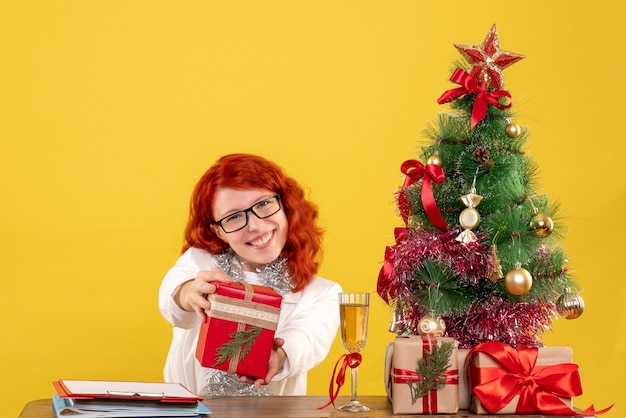 黄色の背景にクリスマスプレゼントとテーブルの後ろに座っている正面図の女性医師