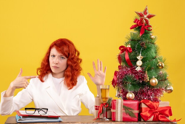 크리스마스 테이블 뒤에 앉아 전면보기 여성 의사 노란색 배경에 선물