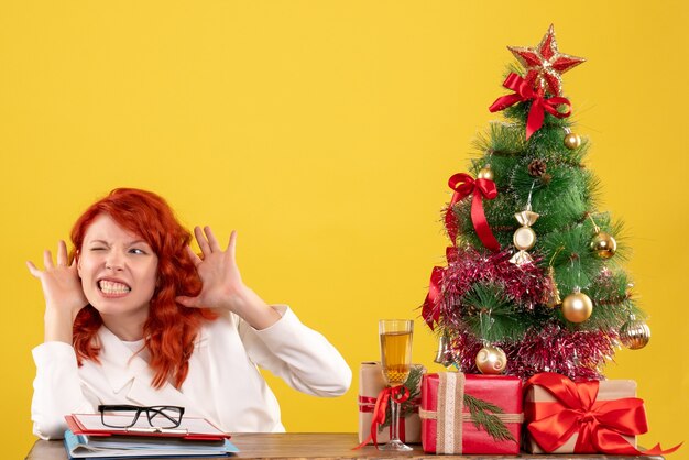 黄色の背景にクリスマスプレゼントとテーブルの後ろに座っている正面図の女性医師