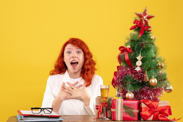 Front view female doctor sitting behind table with christmas presents on yellow background