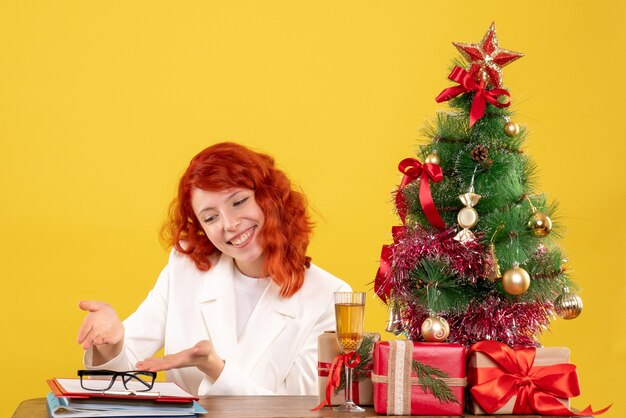 Front view female doctor sitting behind table with christmas presents on yellow background