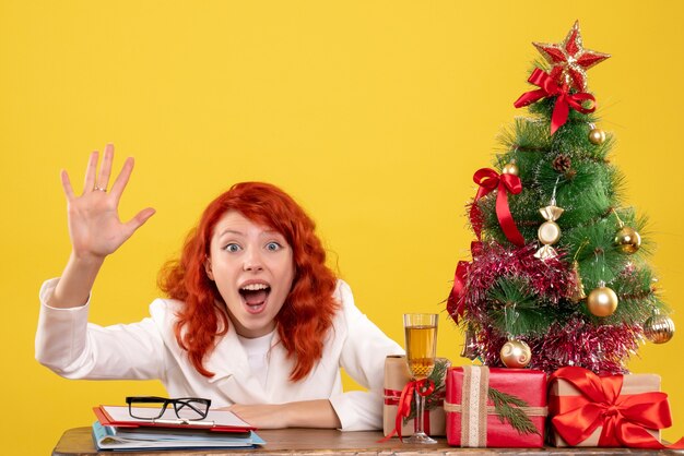 Front view female doctor sitting behind table with christmas presents on yellow background