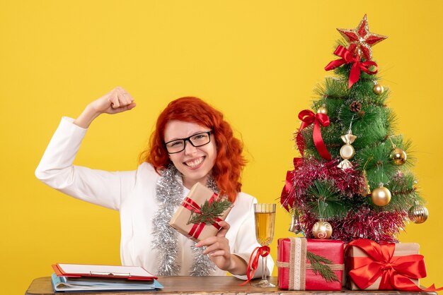 Front view female doctor sitting behind table with christmas presents on yellow desk