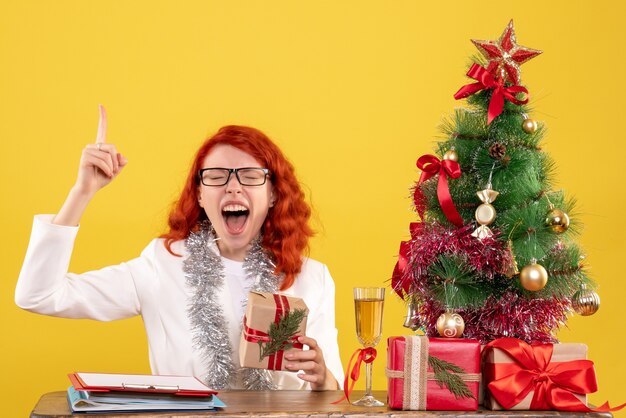Вид спереди женщина-врач сидит за столом с рождественскими подарками на желтом столе
