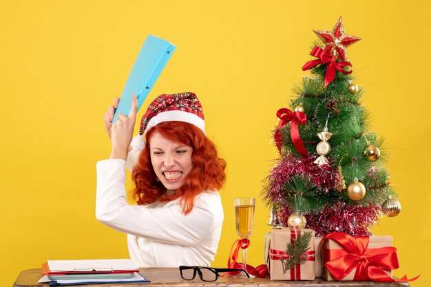 クリスマスツリーとギフトボックスと黄色の背景に怒っている彼女の手でドキュメントとテーブルの後ろに座っている正面図の女性医師