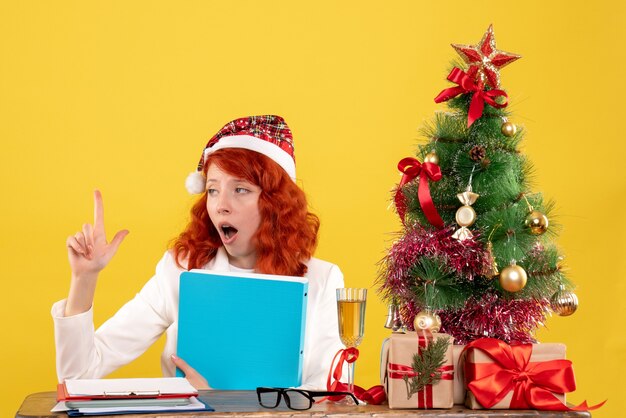 クリスマスツリーとギフトボックスと黄色の背景に彼女の手でドキュメントとテーブルの後ろに座っている正面図の女性医師