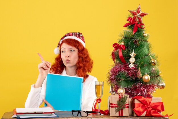 Front view female doctor sitting behind table with documents in her hands on yellow background with christmas tree and gift boxes