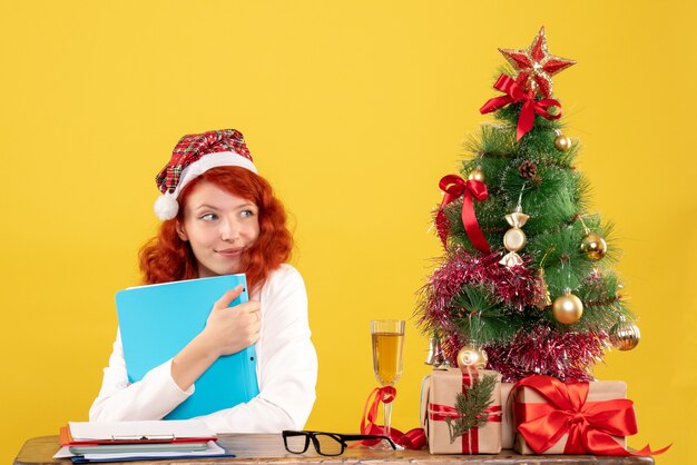 Front view female doctor sitting behind table with documents in her hands on yellow with christmas tree and gift boxes