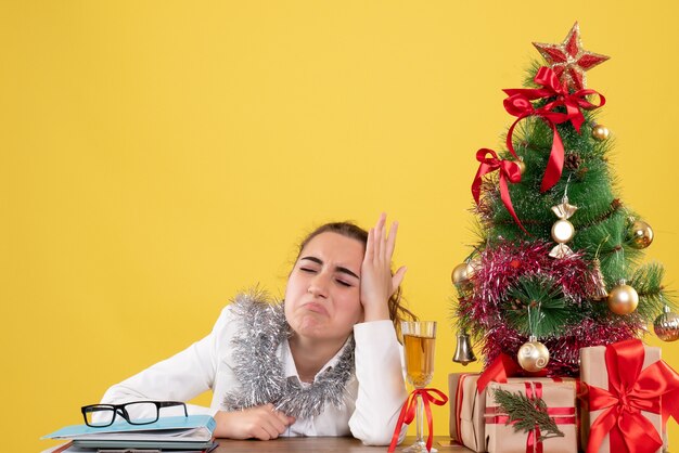 Вид спереди женщина-врач, сидящая за столом с рождественскими подарками и грустным деревом на желтом фоне