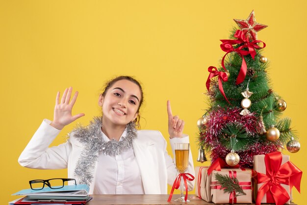 크리스마스 트리와 선물 상자와 노란색 배경에 테이블 뒤에 앉아 전면보기 여성 의사