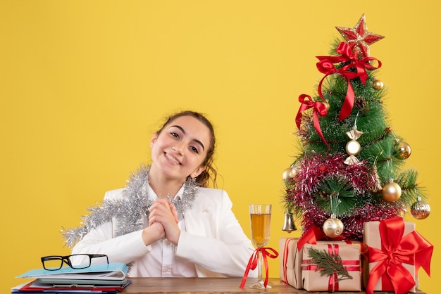 크리스마스 트리와 선물 상자와 노란색 배경에 테이블 뒤에 앉아 전면보기 여성 의사