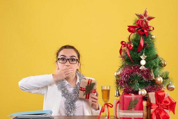 Вид спереди женщина-врач сидит с рождественскими подарками и елкой на желтом фоне h