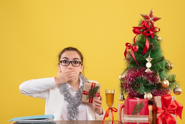 Front view female doctor sitting with christmas presents and tree on yellow background h