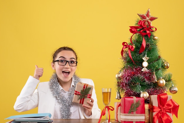 Free photo front view female doctor sitting with christmas presents and tree on a yellow background