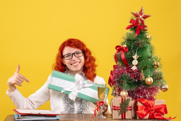 黄色の背景にクリスマスプレゼントと木と一緒に座っている正面図の女性医師