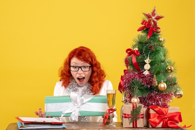 Front view female doctor sitting with christmas presents and tree on yellow background