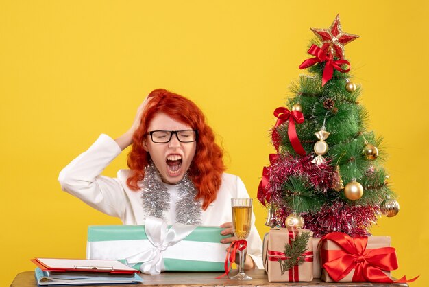 Front view female doctor sitting with christmas presents and tree on yellow background