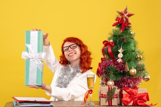 Front view female doctor sitting with christmas presents and tree on yellow background