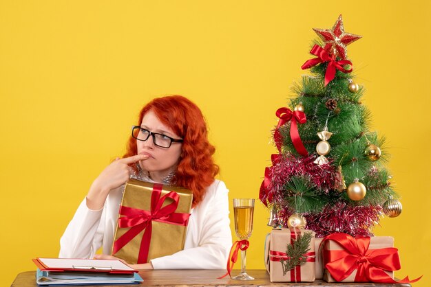黄色の背景にクリスマスプレゼントと座っている正面図の女性医師