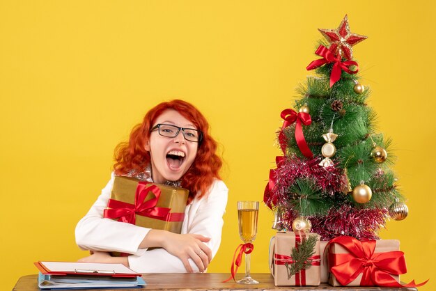 黄色の背景にクリスマスプレゼントと座っている正面図の女性医師